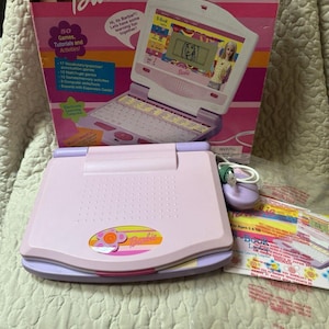 Op de afbeelding: Een Barbie B-Book laptopspeelgoed in een lichtpaarse en roze kleurstelling, inclusief een bijpassende computermuis met snoer. Het speelgoed staat voor de originele kartonnen verpakking, waarop de productnaam en kenmerken staan.
