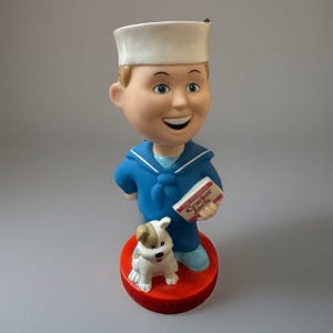 May include: A bobblehead figurine of the Cracker Jack sailor boy wearing a blue uniform and white hat, holding a box labeled The Original Cracker Jack. A small white and brown dog sits at the sailor's feet on a red circular base with text.