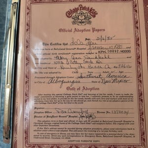 May include: Official adoption papers for a Cabbage Patch Kid, featuring the Cabbage Patch Kids logo. The document includes details like the birth date, adoption date, and the names of the child and adoptive parents. The document is in a blue binder.