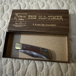 May include: A folding pocket knife with a wood handle and brass bolsters resting inside a brown cardboard box. The box lid features gold text reading SCHRADE Cutlery, THE OLD-TIMER, and A Knife Like Grandad's, with SCHRADE CUTLERY CORP ELLENVILLE, N.Y.