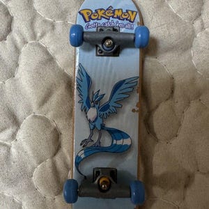 Peut inclure: Un finger skate avec un plateau bleu clair orné d'une illustration du Pokémon Artikodin et du texte Pokémon Gotta catch 'em all! La planche est équipée de trucks gris et de quatre petites roues bleues fixées sous le plateau.