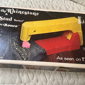 May include: A rectangular cardboard box for The Rhinestone & Stud Setter by Ronco. The box features a yellow and red tool, an illustration of a fabric piece with rhinestones, and the text "The Rhinestone & Stud Setter by Ronco" and "As seen on TV".