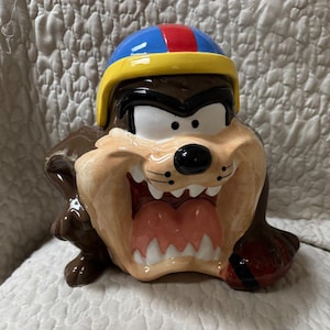 Könnte beinhalten: Eine Keramikfigur des Tasmanischen Teufels, der einen blauen, roten und gelben Football-Helm trägt. Die Figur hat braunes Fell, ein beiges Gesicht, weiße Zähne, eine rosa Zunge und hält einen braunen Football in einer Hand.