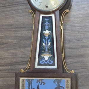 May include: A wooden wall clock featuring a round face with black numerals, topped by a gold eagle finial. The central panel displays a vertical floral motif in blue, white, and gold, while the lower rectangular panel depicts a landscape scene.