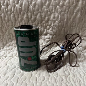 May include: A vintage 7-Up soda can repurposed as an electrical lamp base, featuring the classic green and white branding with the text SEVEN-UP and 12 FLUID OUNCES. A brown electrical cord with a plug is attached to the side of the metal cylinder.