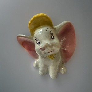 May include: A ceramic wall plaque shaped like a baby elephant with large pink ears, a yellow hat, and a yellow collar. The elephant has a glossy white finish, dark painted eyes with long eyelashes, and a small trunk. The piece is sculpted in relief.