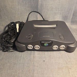Pode incluir: Uma consola de videojogos Nintendo 64 cinzento-carvão com uma ranhura frontal para cartuchos, um botão de ligar, um botão de reiniciar e quatro portas para comandos. Um cabo de alimentação preto com um conetor branco está enrolado à esquerda.