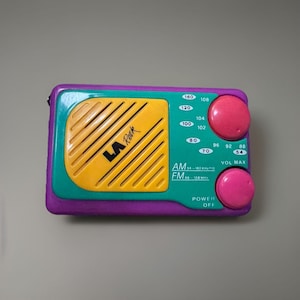 May include: A vintage portable radio with a purple, teal, and yellow color scheme. The radio features a speaker grill with the text "LA Rock", tuning dials, and a power switch. The radio measures approximately 5 inches long and 3 inches wide.