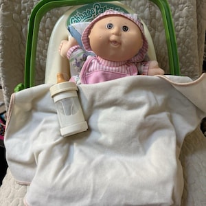 May include: A Cabbage Patch Kids doll with a round face and brown eyes, wearing a pink and white striped bonnet and outfit. The doll sits in a green plastic carrier with a cream-colored blanket draped over it, accompanied by a small toy baby bottle.