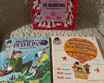Op de afbeelding: Drie vintage Walt Disney platen- en boeksets met 101 Dalmatiërs met een rode hoes, Peter Pan met een blauwe hoes en It's a Small World met een crèmekleurige hoes. Elke vierkante hoes toont kleurrijke illustraties van personages en tekst.