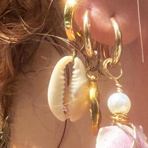 May include: Close-up of gold hoop earrings with seashell, pearl, and gold charm pendants. The earrings are worn on a person's ear, with strands of hair visible in the background. The seashell is cream-colored, and the pearl is white.