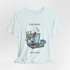 Könnte beinhalten: Hellblaues T-Shirt mit einem Cartoon-Laptop-Motiv. Der Laptop-Bildschirm zeigt herausspringende Werkzeuge, mit dem Text "PLEASE STAND BY..." darüber und "DAD IS FIXING IT" darunter. Eine blaue Tasse ist ebenfalls vorhanden.