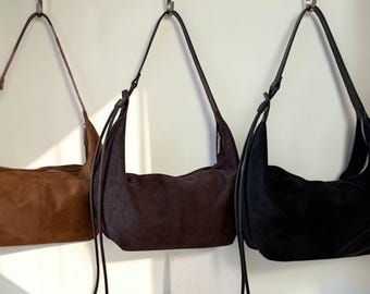 Pode incluir: Três bolsas de ombro de camurça em tons de castanho, castanho escuro e preto penduradas em uma barra de metal. Cada bolsa tem uma alça longa e fina e um fecho de cordão. As bolsas são exibidas contra uma parede branca.