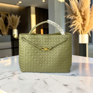 K&ouml;nnte beinhalten: Eine olivgr&uuml;ne, gewebte Handtasche mit einem oberen Griff und einem goldfarbenen Verschluss. Die Tasche hat ein geflochtenes Detail und einen wei&szlig;en Griff. Die Tasche ist rechteckig und hat eine strukturierte Form.