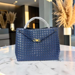 K&ouml;nnte beinhalten: Eine marineblaue, gewebte Handtasche mit wei&szlig;em Griff. Die Tasche hat einen goldfarbenen Verschluss und einen Klappenverschluss. Die Tasche steht auf einer wei&szlig;en Oberfl&auml;che.