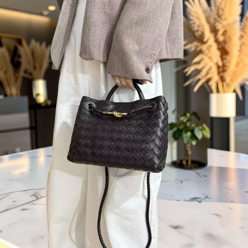 K&ouml;nnte beinhalten: Dunkelbraune Handtasche aus geflochtenem Leder mit einem oberen Griff und einem goldfarbenen Verschluss. Die Tasche hat einen geflochtenen Kordelzugverschluss und einen abnehmbaren Schulterriemen. Die Tasche wird von einer Person gehalten, die einen Blazer und eine wei&szlig;e Hose tr&auml;gt.