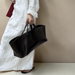 Peut inclure: Un grand sac fourre-tout marron foncé avec deux poignées est tenu par une personne vêtue d'une robe blanche. Le sac semble être en cuir ou dans une matière similaire. L'arrière-plan est de couleur neutre unie.