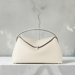 Puede incluir: Bolso de mano color crema con asa curva y cierre plateado. El bolso tiene una forma estructurada con ribete negro. El bolso está sobre una superficie blanca contra un fondo gris texturizado.