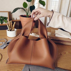Peut inclure: Un sac à main en cuir marron avec un design doux et froncé. Le sac a une base structurée et une poignée incurvée. L'image montre le sac sur une surface en bois, avec une plante et d'autres objets en arrière-plan.