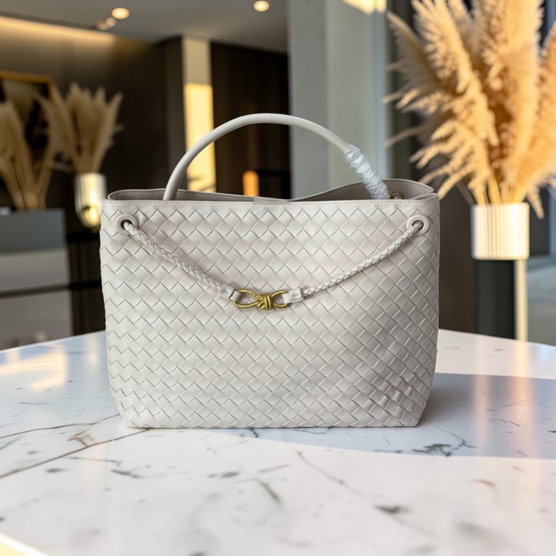 K&ouml;nnte beinhalten: Eine hellgraue Handtasche aus gewebtem Leder mit einem oberen Griff und einem geflochtenen Riemenverschluss. Die Tasche hat einen goldfarbenen Verschluss und ein strukturiertes Design. Der Innenraum ist sichtbar, und die Tasche scheint leer zu sein.