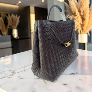K&ouml;nnte beinhalten: Dunkelbraune Handtasche aus geflochtenem Leder mit einem oberen Griff und einem geflochtenen Riemen mit einem goldfarbenen Verschluss. Die Tasche hat eine strukturierte Form und ein ger&auml;umiges Innenfach.