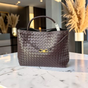 K&ouml;nnte beinhalten: Dunkelbraune, gewebte Ledertasche mit oberem Griff und geflochtenen Schultergurten. Die Tasche hat einen goldfarbenen Verschluss und ein ger&auml;umiges Innenfach. Die Tasche steht auf einer wei&szlig;en Marmoroberfl&auml;che.