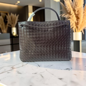 K&ouml;nnte beinhalten: Dunkelgraue, gewebte Ledertasche mit oberem Griff und Kettendetail. Die Tasche hat eine rechteckige Form und ein geflochtenes Kettendetail am oberen Rand. Der Griff hat eine passende dunkelgraue Farbe.