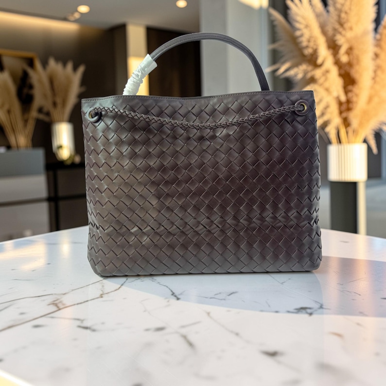 K&ouml;nnte beinhalten: Dunkelgraue, gewebte Ledertasche mit oberem Griff und Kettendetail. Die Tasche hat eine rechteckige Form und ein geflochtenes Kettendetail am oberen Rand. Der Griff hat eine passende dunkelgraue Farbe.