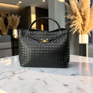 K&ouml;nnte beinhalten: Schwarze Tote Bag aus geflochtenem Leder mit einem oberen Griff und einem goldfarbenen Verschluss. Die Tasche hat ein geflochtenes Detail auf der Vorderseite und eine strukturierte Form. Die Tasche steht auf einer wei&szlig;en Marmoroberfl&auml;che.