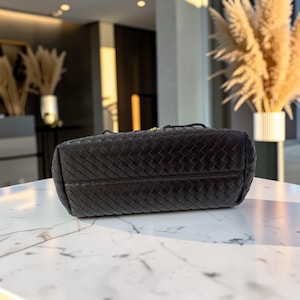 K&ouml;nnte beinhalten: Eine schwarze Handtasche aus geflochtenem Leder mit rechteckiger Form. Die Tasche hat ein Korbgeflechtmuster und einen strukturierten Boden. Das Bild ist aus einem niedrigen Winkel aufgenommen, der die Textur und das Design der Tasche hervorhebt. Der Hintergrund ist verschwommen.