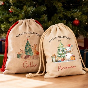 May include: Two beige drawstring gift bags with red and beige drawstrings. One bag features a Christmas tree, fox, and bunny illustration with the name "Callie." The other bag has a snowman and Christmas tree illustration with the name "Arianna." Both bags say "Special Delivery for."
