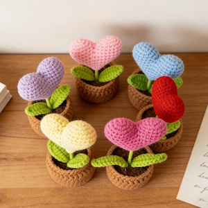 Puede incluir: Una colección de flores hechas a mano en forma de corazón de ganchillo en pequeñas macetas marrones. Las flores son de varios colores, incluyendo rosa, morado, amarillo, rojo y azul, con hojas verdes. La disposición está sobre una superficie de madera.