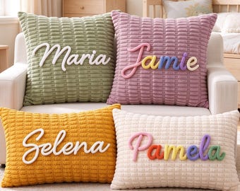 Può includere: Quattro cuscini quadrati e rettangolari con un motivo a griglia testurizzato. Il cuscino verde oliva presenta il nome bianco Maria, il cuscino malva ha il nome multicolore Jamie, il cuscino oro ha il nome bianco Selena e il cuscino crema ha il nome multicolore Pamela.