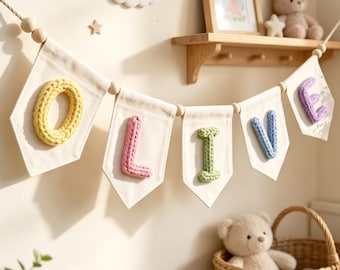 May include: A decorative banner featuring five off-white fabric pennants strung on a rope with wooden beads. Each pennant displays a single, chunky, knit-textured letter in pastel colors, spelling out the name OLIVE in yellow, pink, green, blue, and purple.