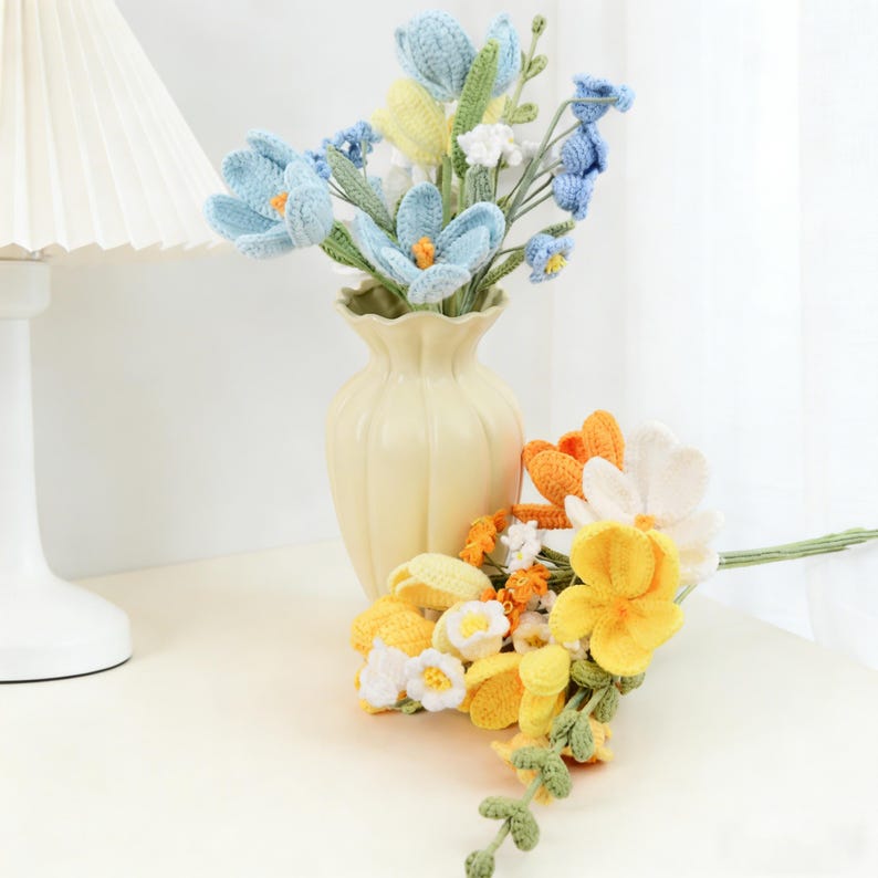 以下が含まれることがあります： クリーム色の花瓶には、青、黄、白の色合いのかぎ針編みの花のブーケが入っています。2つ目の黄、オレンジ、白のかぎ針編みの花のブーケが花瓶の前にあります。花は白い表面に配置されています。