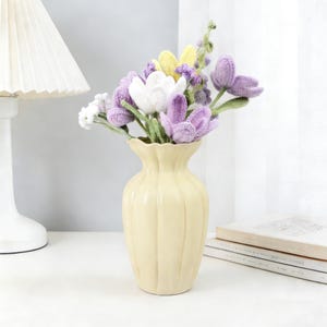 以下が含まれることがあります： クリーム色の溝のあるデザインの花瓶には、手作りのかぎ針編みの花のブーケが入っています。花には、白い花、黄色い花、紫の花と緑の茎が含まれています。花瓶は、ランプと本に隣接する白い表面に置かれています。