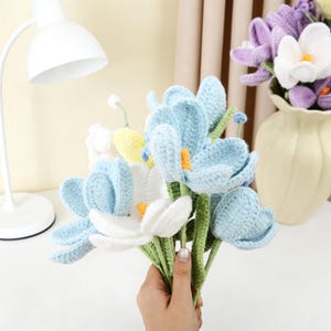 以下が含まれることがあります： 水色と白を基調とした手編みの花のブーケ。黄色とオレンジのアクセントが施されています。花は緑色の茎を持ち、手で持たれています。背景には紫と白の手編みの花が入った花瓶があります。