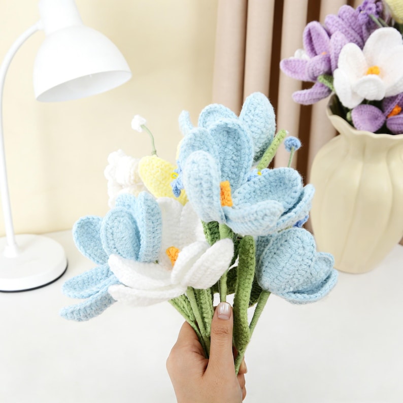 以下が含まれることがあります： 水色と白を基調とした手編みの花のブーケ。黄色とオレンジのアクセントが施されています。花は緑色の茎を持ち、手で持たれています。背景には紫と白の手編みの花が入った花瓶があります。