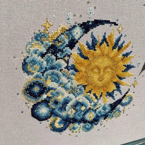 Puede incluir: Arte de punto de cruz con un diseño celestial. Un sol dorado con una cara sonriente está rodeado por una luna creciente y nubes azules y doradas. La obra de arte está adornada con pequeñas cuentas y estrellas brillantes.