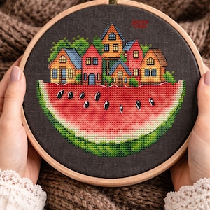 May include: Cross-stitch embroidery in a wooden hoop. The design features a village of colorful houses atop a watermelon slice. The houses have blue, red, and yellow roofs. The watermelon has black seeds and green rind.