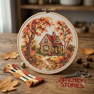 May include: A circular wooden embroidery hoop containing a cross-stitch design of a house surrounded by autumn trees in shades of orange, red, and yellow. The scene includes a path leading to the house, with embroidery floss, a thimble, and scissors.