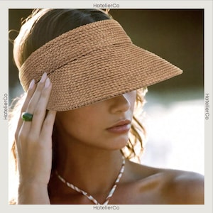 May include: A light brown woven straw visor with a wide brim. The visor has a band to fit around the head. The image also shows a green ring and a white and brown necklace. The text "HatellierCo" is visible.