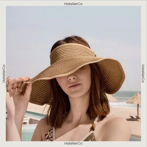 May include: A wide-brimmed, woven straw visor in a light brown hue. The visor has a curved brim and a band that sits atop the head. The image is set against a beach backdrop with the text "HatelierCo".