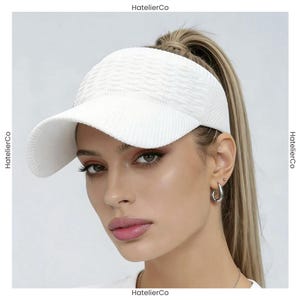May include: A white visor with a textured, wavy pattern on the crown and a ribbed brim. The visor features a structured design with a curved bill, providing a clean and simple look. The brand name HatelierCo is visible on the edges of the image.