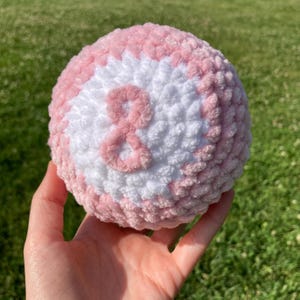 Op de afbeelding: Een handgemaakte, ronde, gehaakte bal in roze en witte tinten. Het nummer "8" is in roze op het witte gedeelte gestikt. De bal wordt in een hand gehouden tegen een vage groene achtergrond.