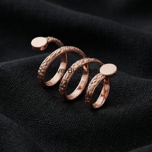 Op de afbeelding: Een roségouden ring met een uniek spiraalontwerp. De ring heeft bloemengravures langs de band en twee cirkelvormige accenten. De ring wordt getoond op een donkere, getextureerde ondergrond.