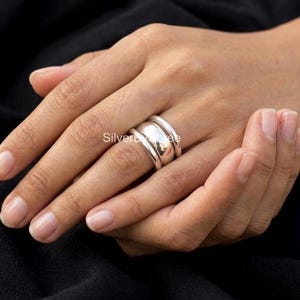 Puede incluir: Un anillo de plata pulida con un diseño de banda ancha, lisa y redondeada. El anillo tiene un acabado metálico de alto brillo y un perfil grueso y sustancial que envuelve el dedo, creando una apariencia de superficie limpia y reflectante.