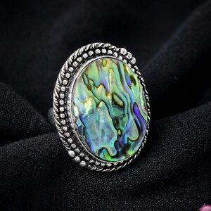 Può includere: Un anello ovale con un grande intarsio in conchiglia di abalone iridescente con motivi vorticosi verdi, blu e viola. La pietra è incastonata in una montatura in metallo argentato ossidato e testurizzato con un bordo a perline.
