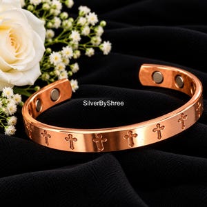 May include: A copper-toned metal cuff bracelet featuring a repeating engraved cross pattern along the outer band. The interior of the open-ended bracelet displays two circular magnetic inserts at each terminal end. The text SilverByShree is visible.