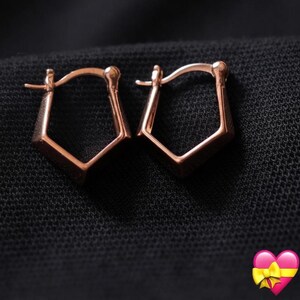 Puede incluir: Un par de pendientes de aro en oro rosa con una forma pentagonal distintiva. Cada pendiente está construido con un mecanismo de poste articulado en la parte superior, creando una silueta geométrica con líneas angulares y un acabado pulido.